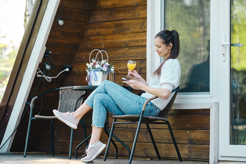 Woman enjoys warm weather sitting on terrace of wooden cabin with smartphone in nature